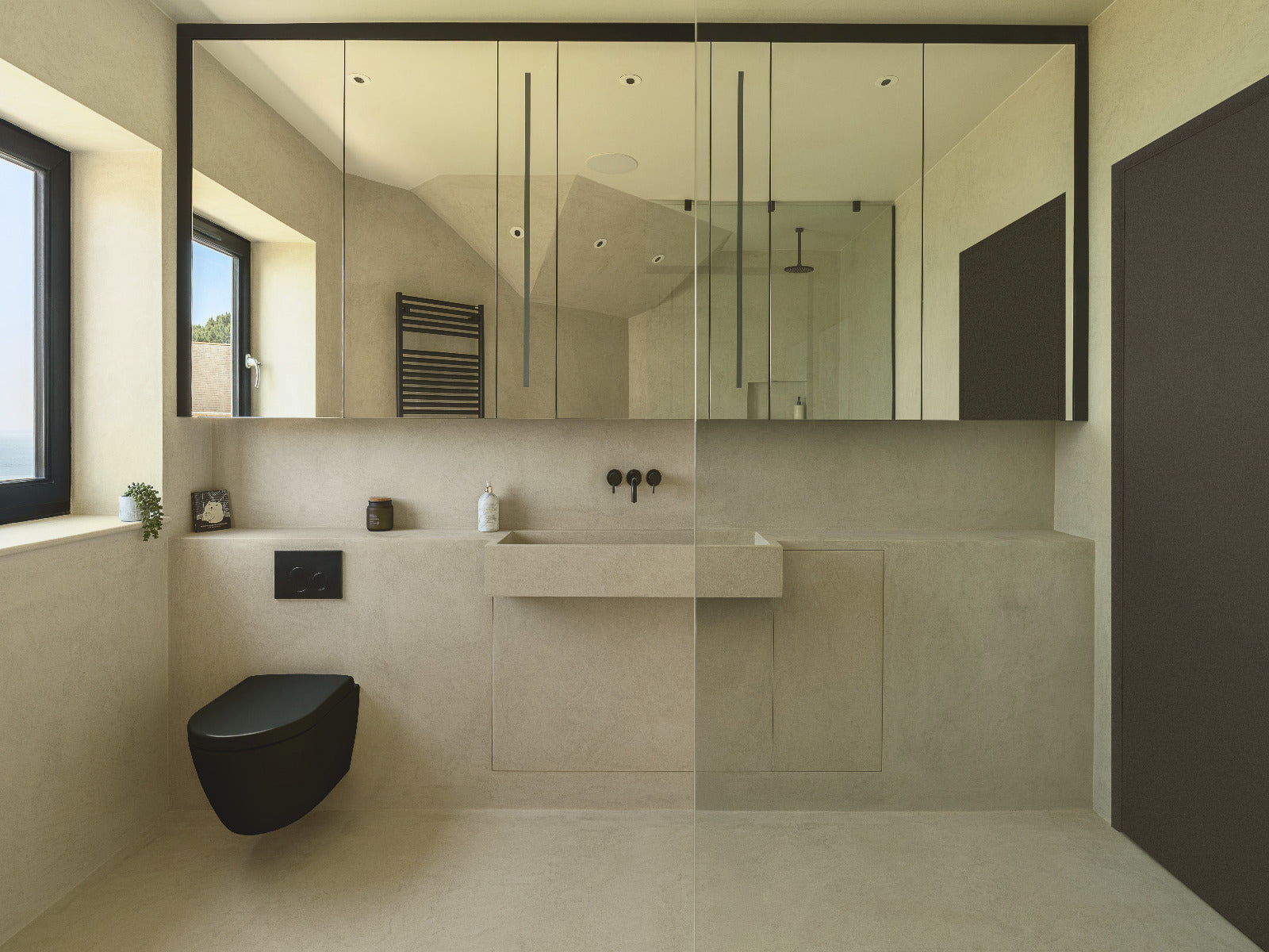 Modern minimalist bathroom interior with a wall-mounted sink, large mirror, black wall-mounted toilet, beige stone tiles, and a dark brown door on the right.