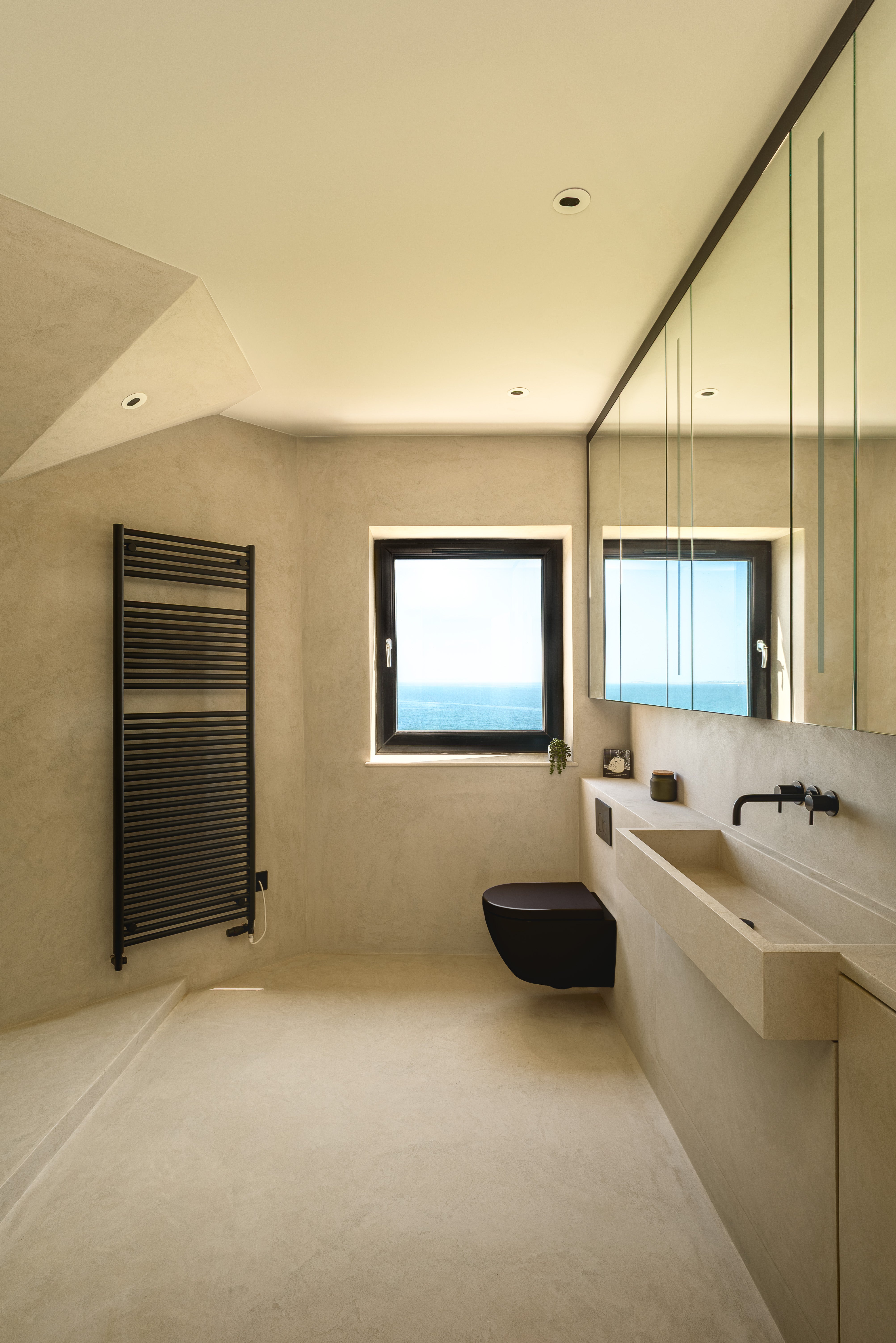 Modern minimalist bathroom with beige plaster walls and floor, a wall-mounted sink, black faucet, black toilet, black heated towel rail, and a large mirror above the sink.