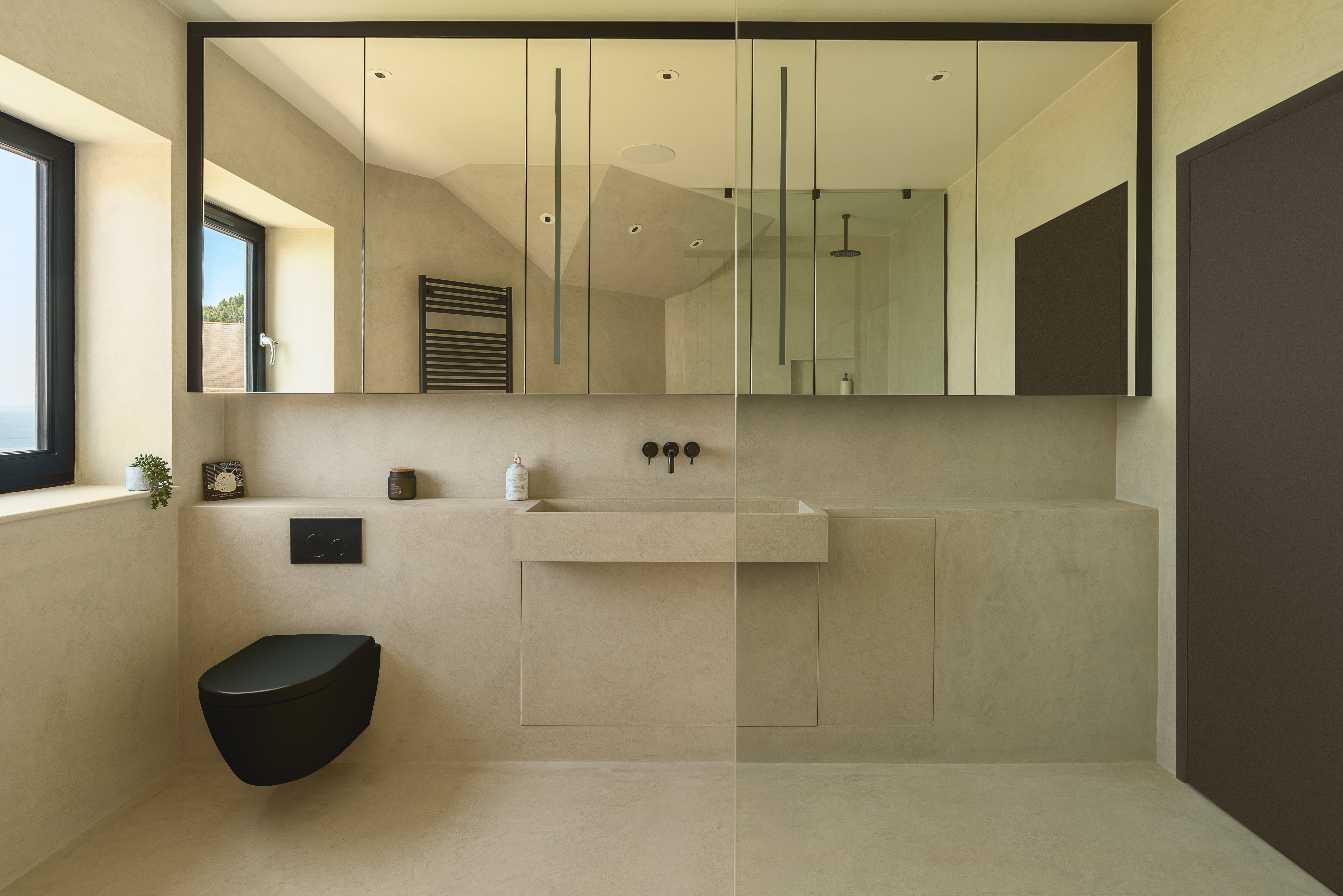 Modern minimalist bathroom interior with a wall-mounted sink, large mirror, black wall-mounted toilet, beige stone tiles, and a dark brown door on the right.