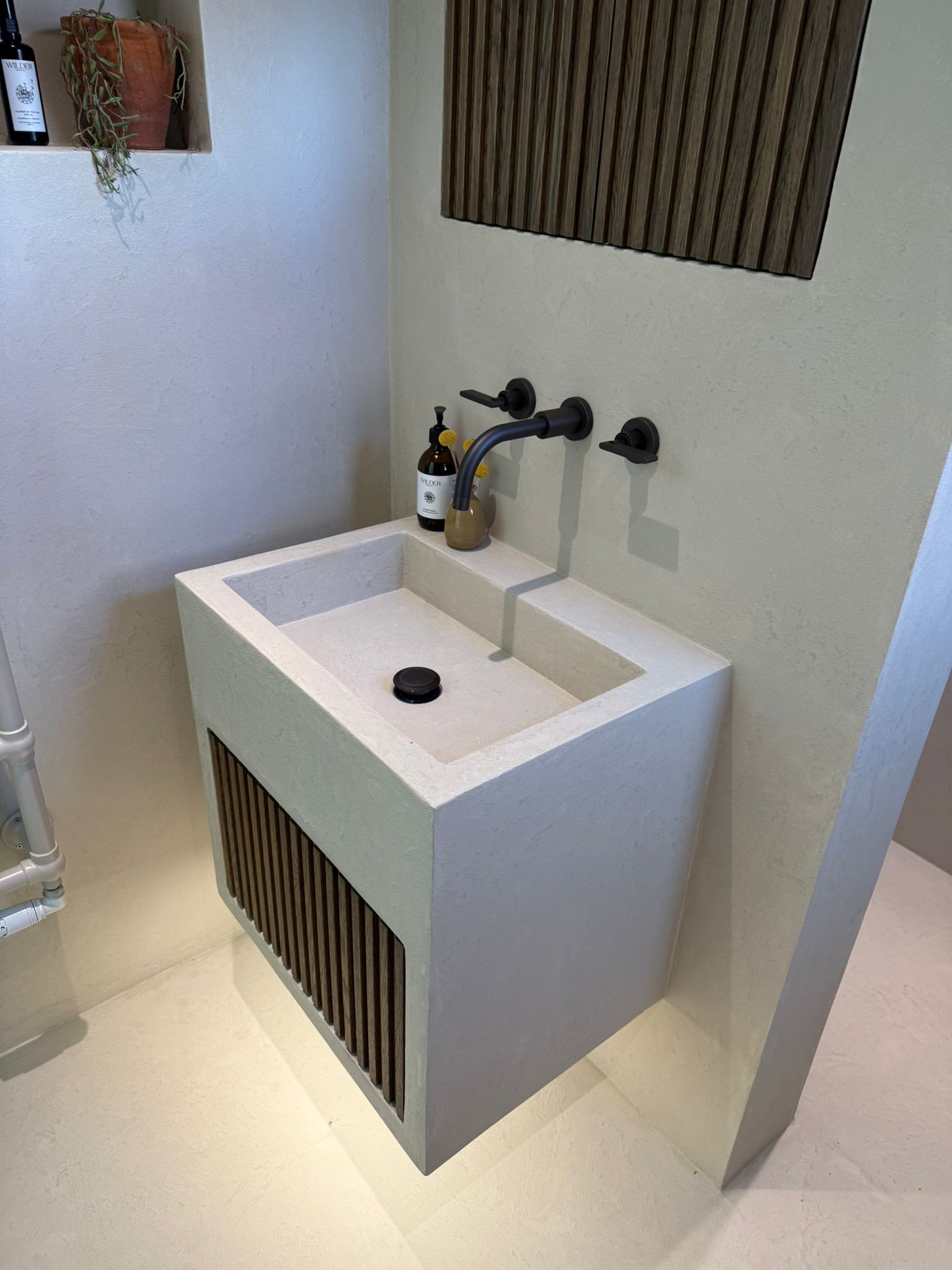 A modern floating bathroom sink with a rectangular basin, dark wood slat accents on the front and wall, matte black faucet and handles, and a light beige wall background.