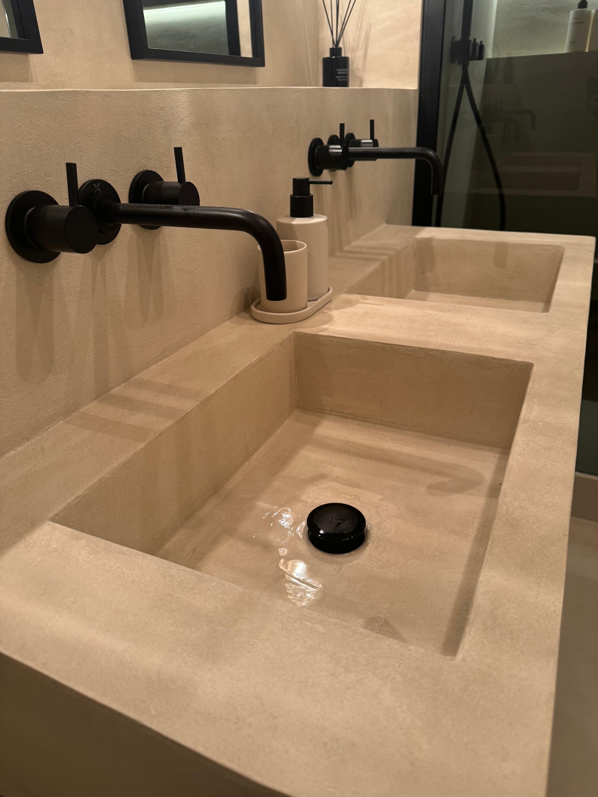 A modern double-sink bathroom vanity featuring a beige stone countertop with two recessed rectangular basins, matte black wall-mounted faucets, and a matching black drain.
